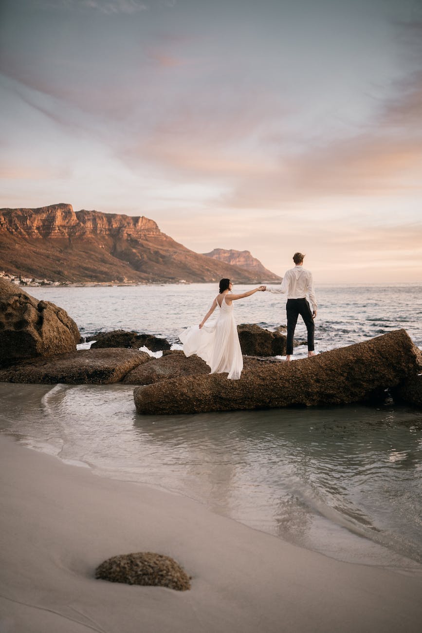 a bride and groom standing on a rock formation at the beach while holding hands